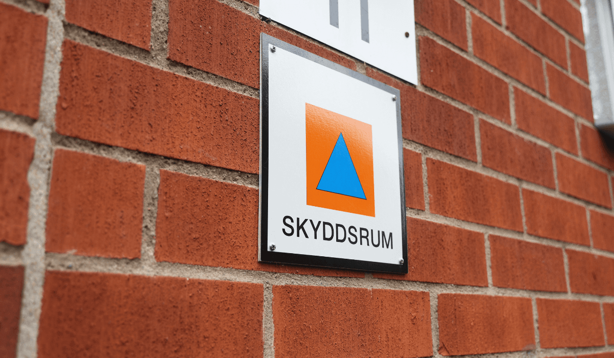 Skylt med skyddsrumssymbol monterad på en fasad av rött tegel.